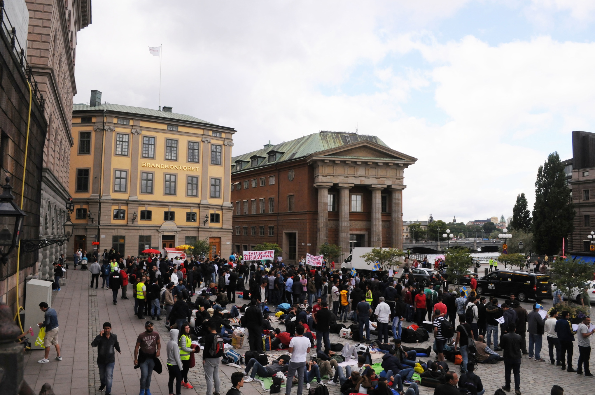 Många av dem som samlats på Mynttorget håller i banderoller och skyltar riktade mot riksdagen; flera står i grupp och diskuterar; andra ligger på liggunderlag på marken och vilar.