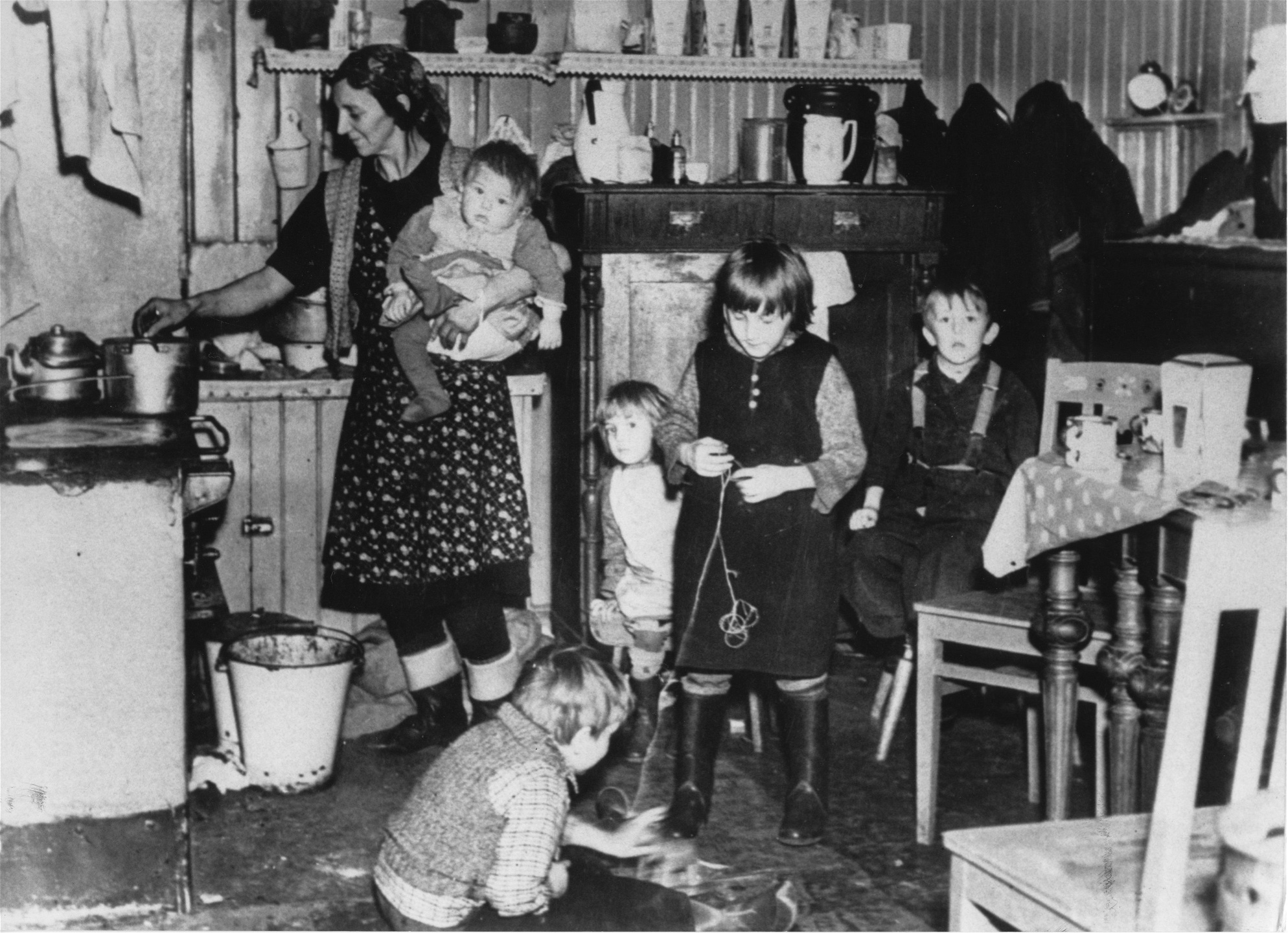 Arbetarfamilj i Stockholm på 1940-talet. En kvinna lagar mat på vedspisen, med ett barn på armen och fyra som leker på köksgolvet.