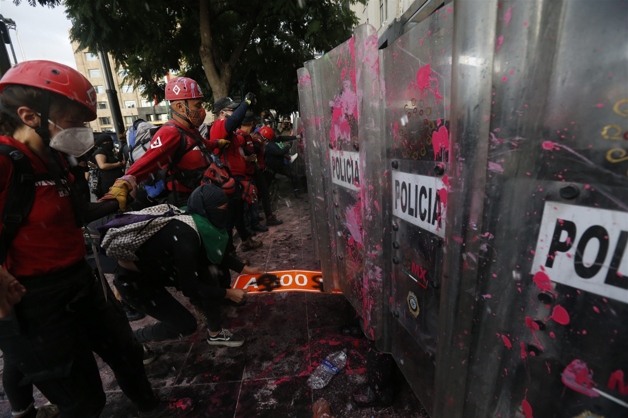 Rödklädda personer med hjälp står mellan demonstranter och poliser med plexiglassköldar som stänkts ned med rosa färg.