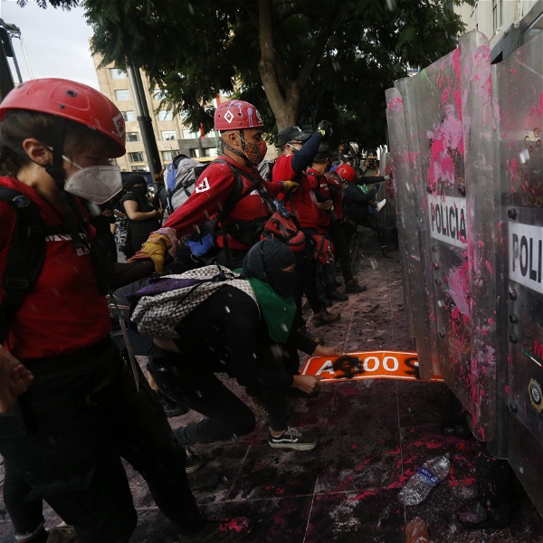 Rödklädda personer med hjälp står mellan demonstranter och poliser med plexiglassköldar som stänkts ned med rosa färg.
