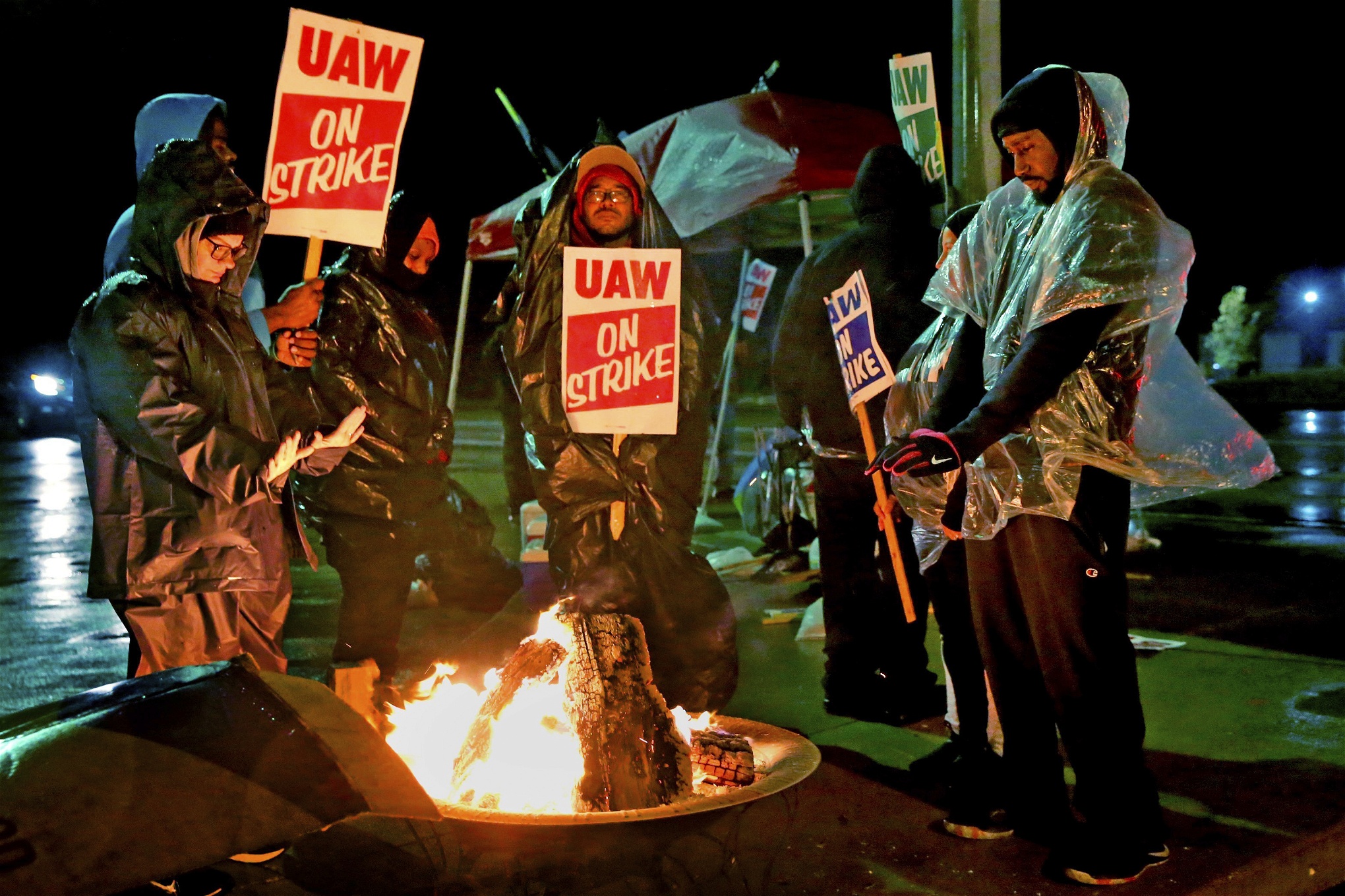 Strejkande bilfabriksarbetare värmer sig runt en brasa. Det är kväll eller natt och de håller skyltar med texten UAW on strike.