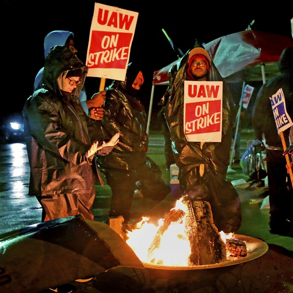 Strejkande bilfabriksarbetare värmer sig runt en brasa. Det är kväll eller natt och de håller skyltar med texten UAW on strike.