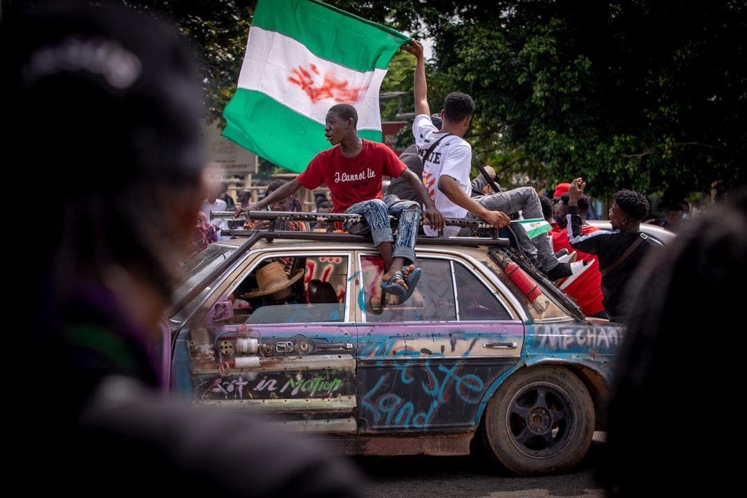 Två unga män sitter på ett biltak och håller i en grönvit flagga, Nigerias flagga. På gatan är fult av folk och bilen är nedklottrad med slagord.
