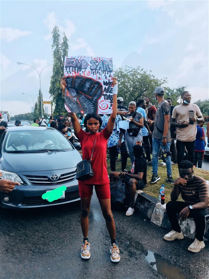 Octavia, ståendes på en gata fylld med demonstranter, håller upp ett plakat med en knuten näve och texten End SARS.