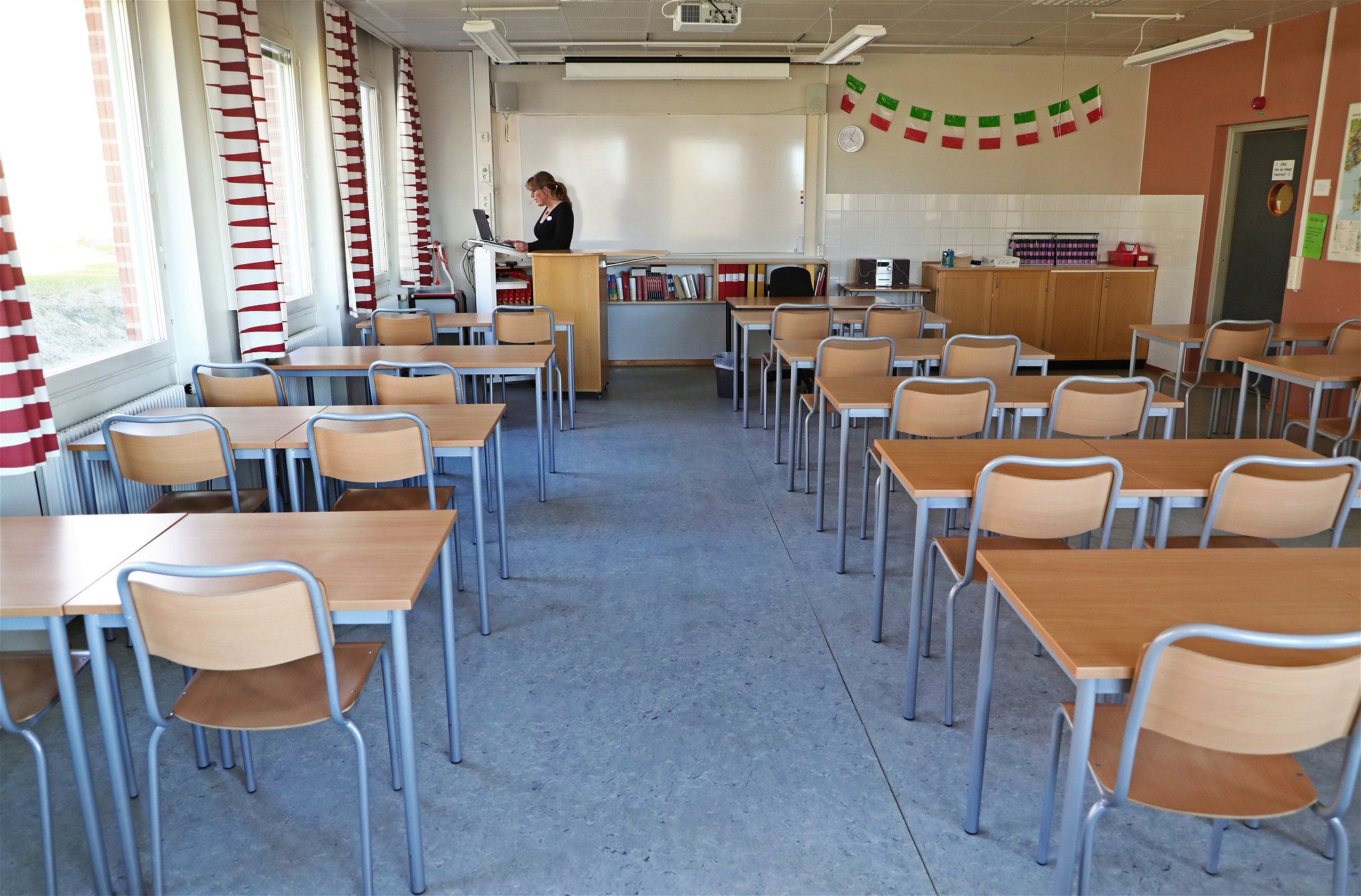 En lärare ensam framför en dator i ett klassrum där bänkarna står tomma.