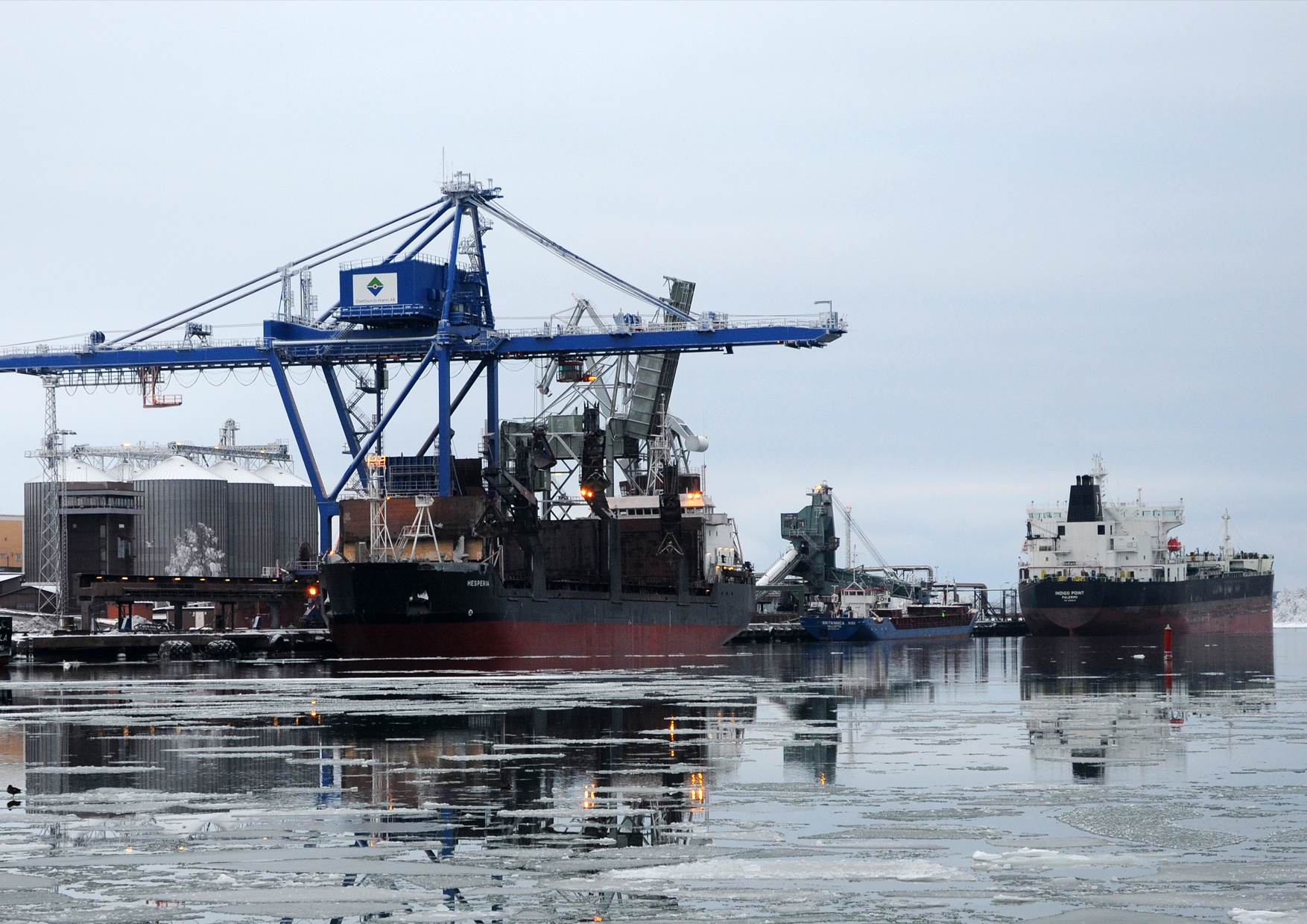 Oxelösunds hamn i januariväder. Bilden visar en blå kran och tre skepp vid kaj