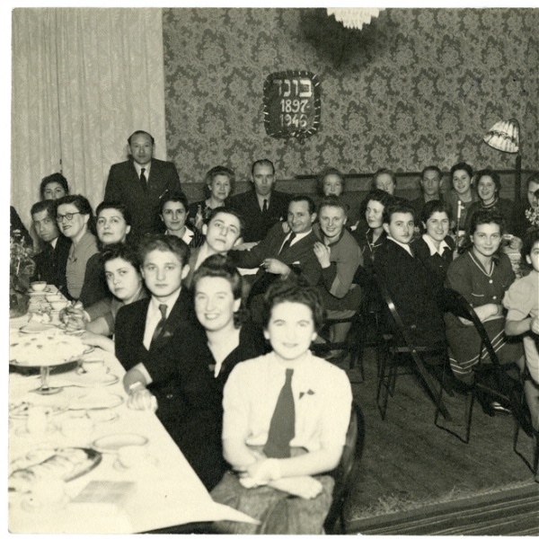 Svartvitt fotografi där en mängd människor sitter samlade kring två bord och tittar in i kameran.