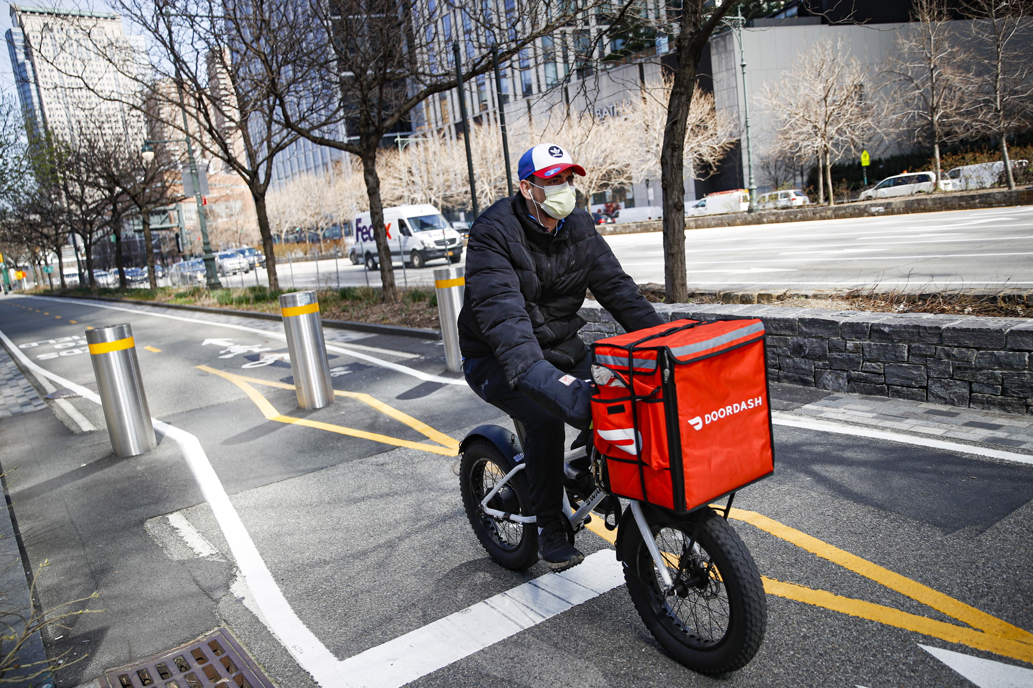 Cykelbud med en röd matförvaringslåda på styret, keps och munskydd, cyklar på en gata.