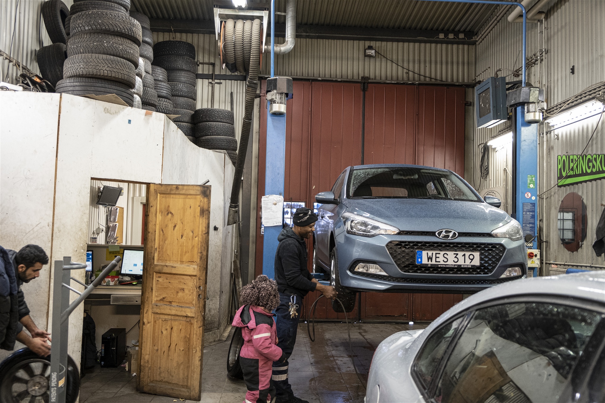 David fäster ett hjul på en upphissad bil i verkstaden. Hans äldsta dotter tittar på i rosa overall. En kollega rullar in ytterligare ett hjul.