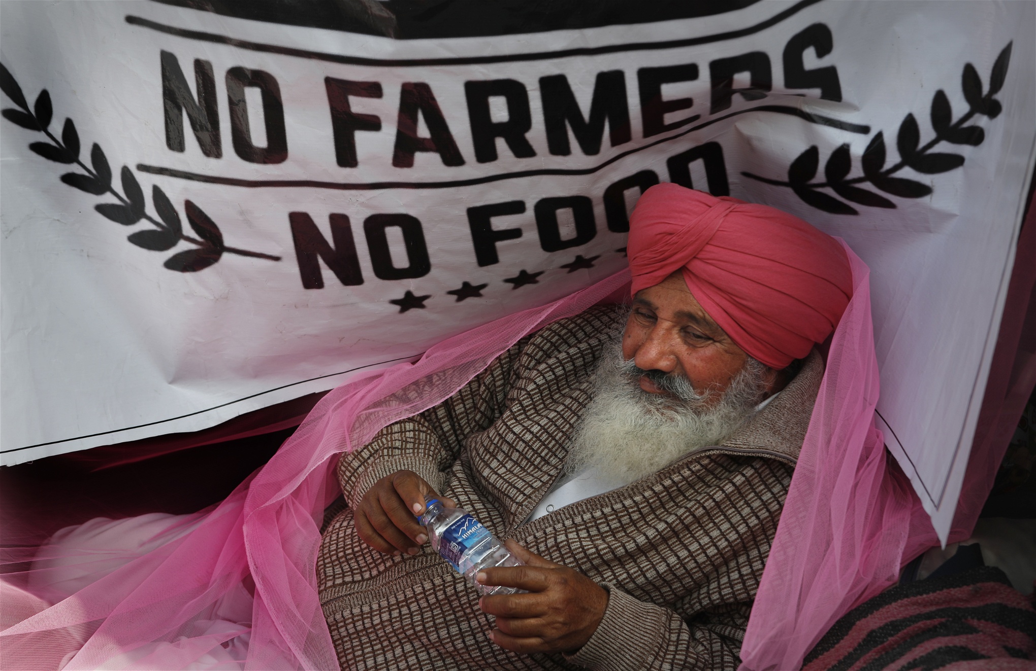 En bonde med en hallonröd turban och en rosa sjal över axlarna lutar sig framför en banderoll med texten No farmers no food.