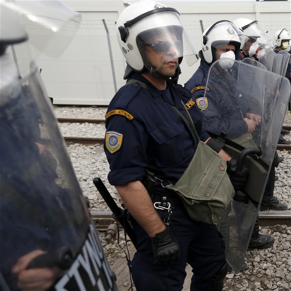 Poliser med vita hjälmar och plexiglassköldar står uppradade längs ett tågspår. En pojke sitter med nedböjt huvud framför dem.