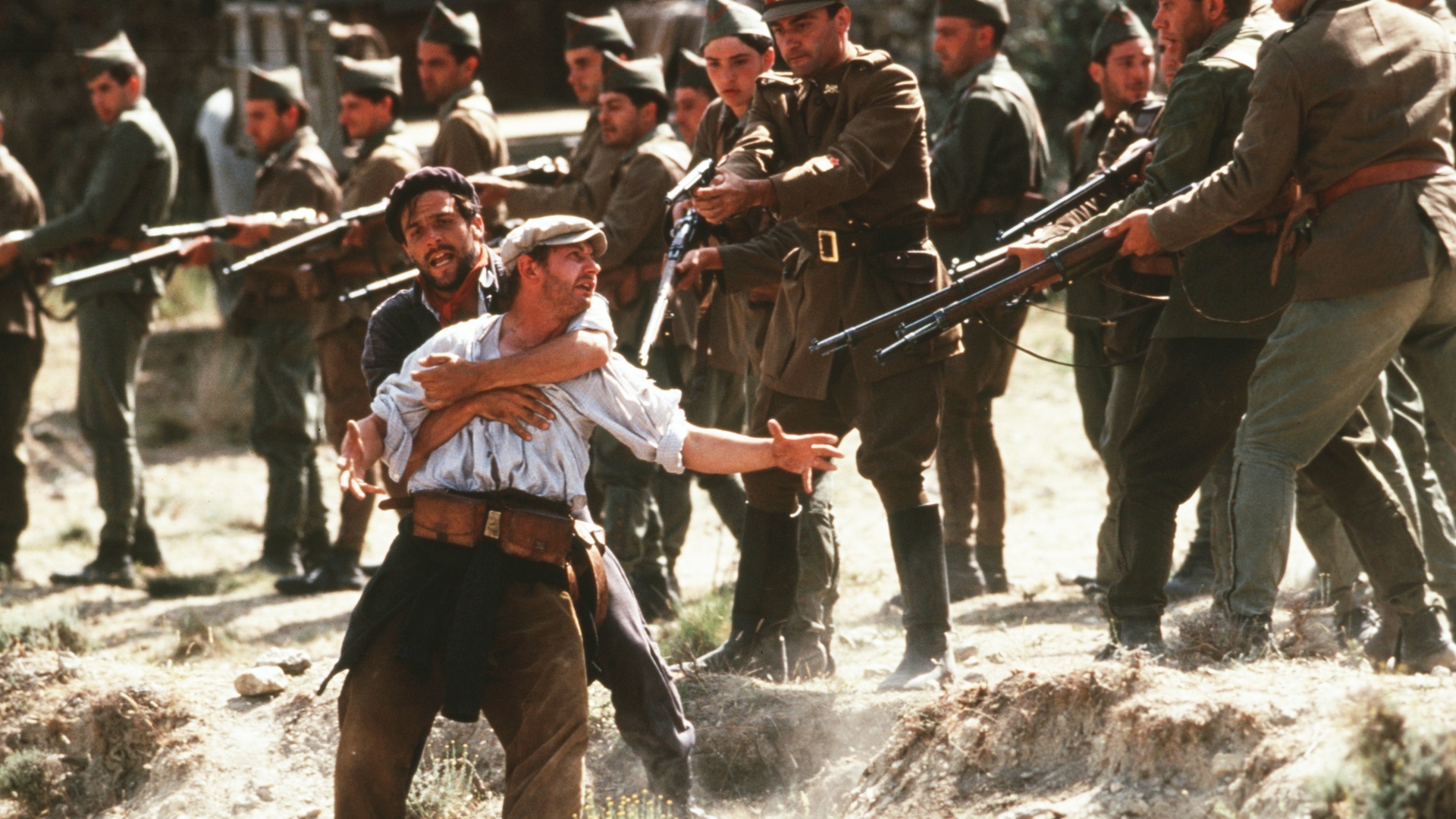 Stillbild från filmen Land och frihet. En man med sjal runt halsen håller om en man med vit skjorta. Soldater i uniformer pekar sina gevär mot dem.