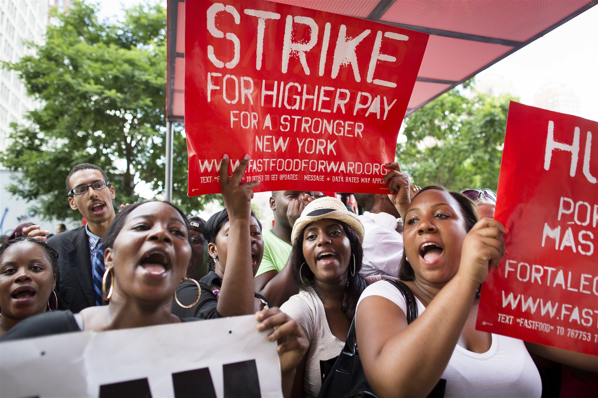 Mestadels afroamerikanska kvinnor i en demonstration. Röda skyltar med texten Strike for higher pay syns.  