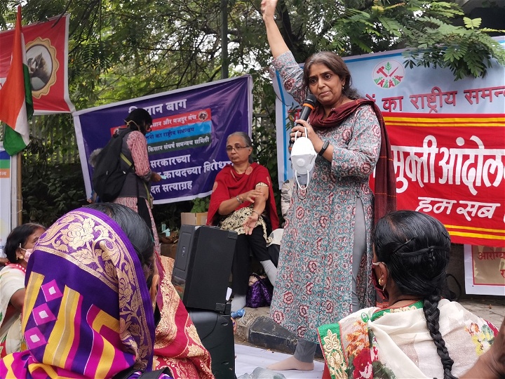 Seema står på en upphöjning, framför plakat och håller ett tal. Ena handen höjer hon i luften.