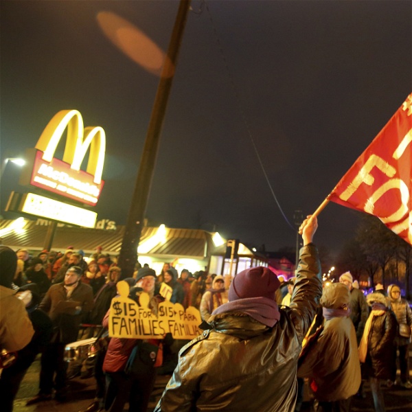 Demonstration framför ett mcdonalds. Det är kväll och mörkt och en demonstrant håller en röd flagga med texten Fight for 15. McDonalds-M:et syns i bakgrunden.