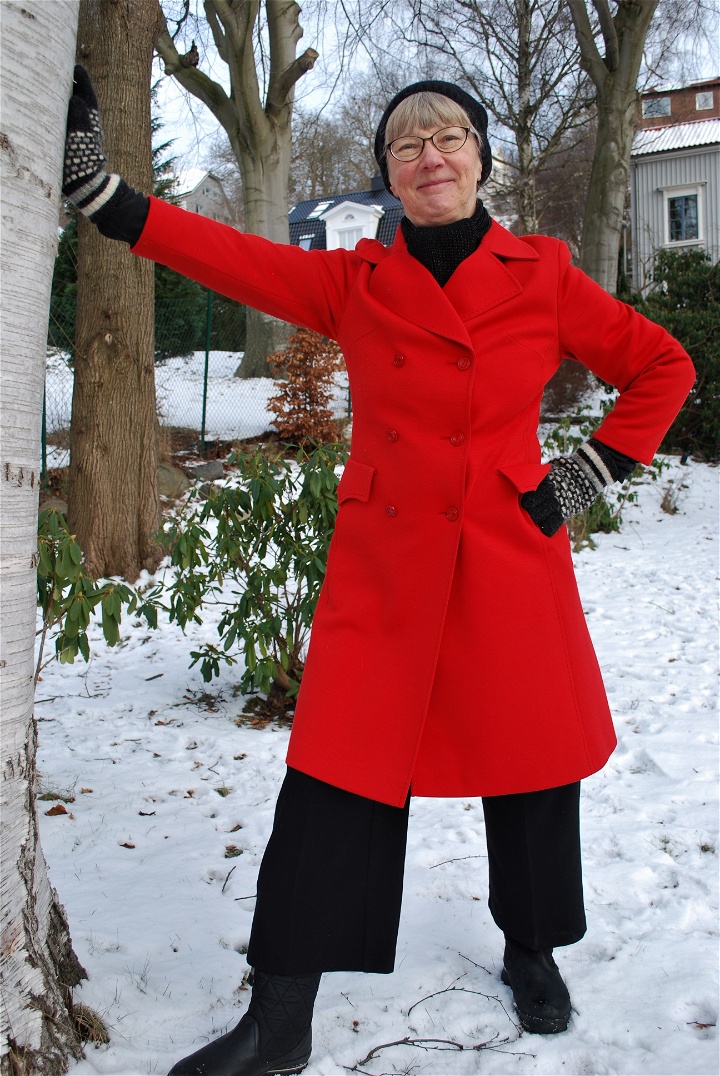 Susanne Fransson, utomhus med armen lutad mot ett träd och andra handen i sidan. Hon har en röd kappa, mössa och vantar. 