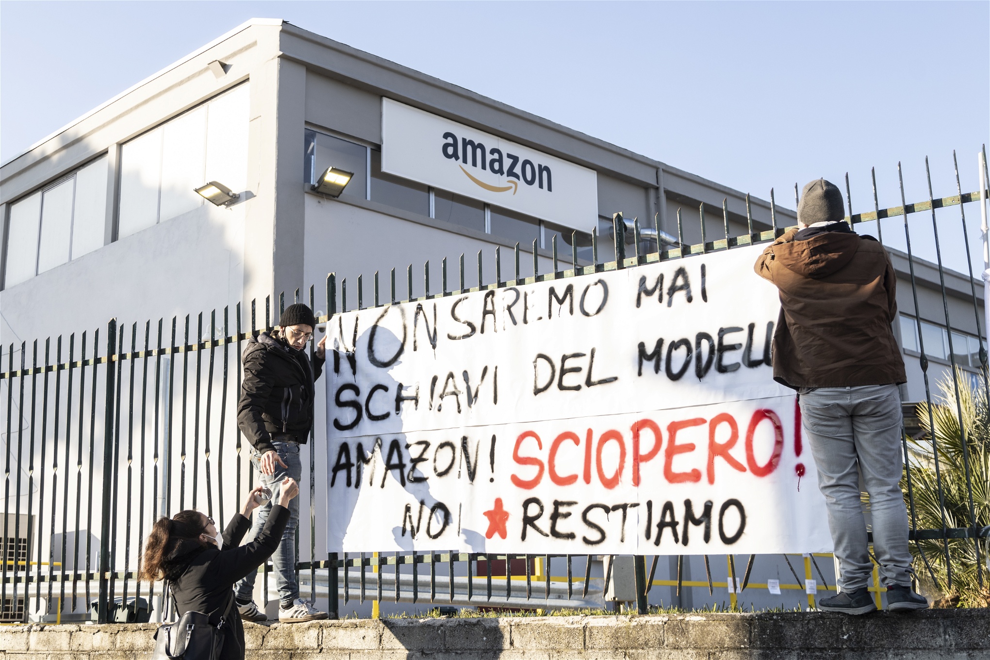 Foto: Valerio NicolosiStrejkande arbetare sätter upp en banderoll på grindarna till Amazon med texten "vi kommer aldrig att vara slavar åt Amazons modell."