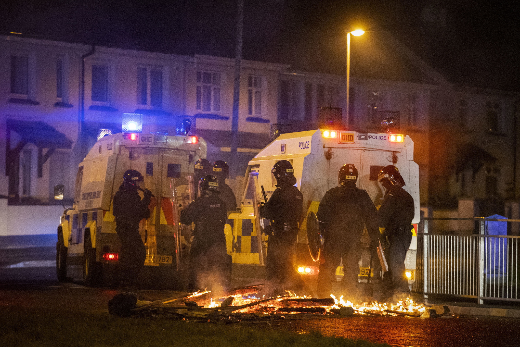 Kravallklädda poliser på en kvällsmörk gata. På markan ligger brinnande plankor.