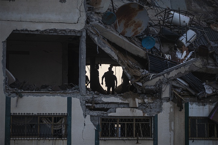 Bombat hus med hål i fasaden och infallet tak. Inne i huset syns människor som söker efter överlevande.