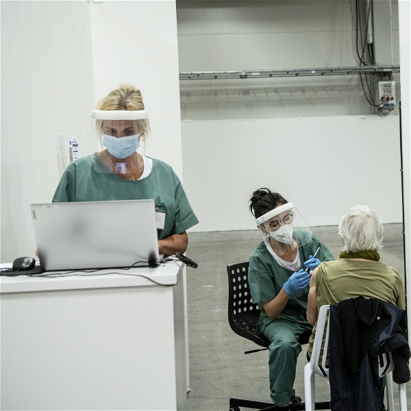 Sjuksköterska står vid en dator, iklädd munskydd och visir. Bakom henne sitter en annan sjuksköterska och vaccinerar en äldre person.