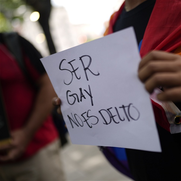 En person med en regnbågsflagga över axlarna håller en skylt med texten: "Att vara gay är inte ett brott", på spanska.