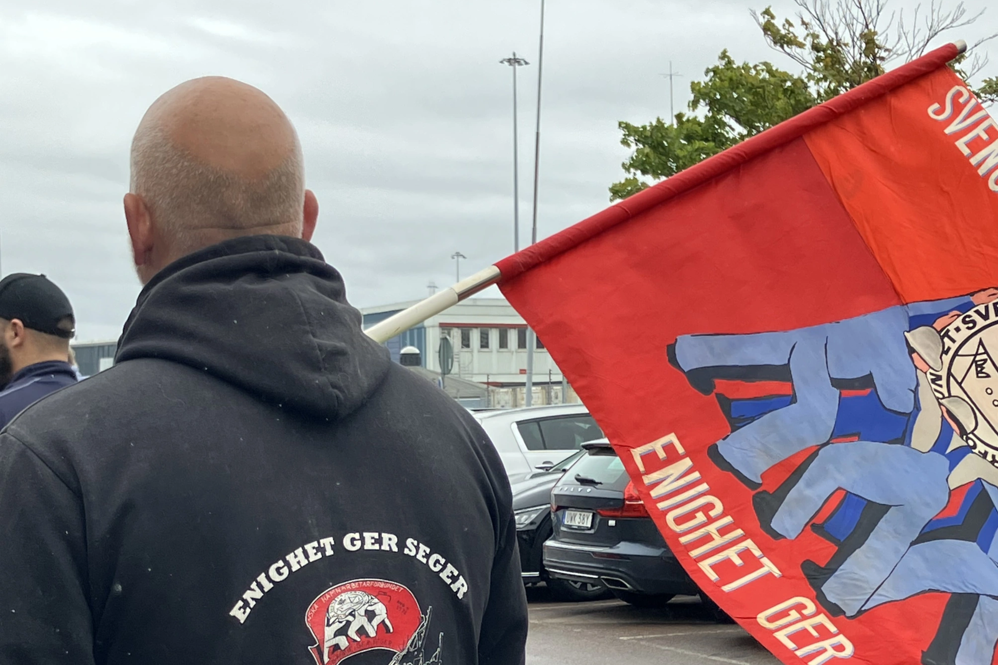 Hamnarbetare i förgrunden under hamnstrejken 2025. På tröjan står det: Enighet ger styrka. Flagga från Hamnarbetarförbundet i bakgrunden