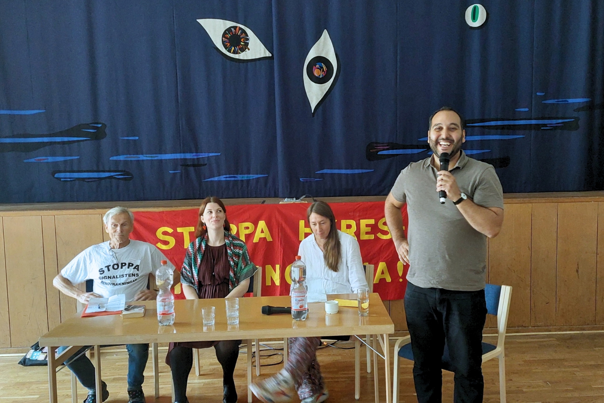 Syntolkning: Foto på en presentation under Bostadsvrålet 2025. Tre personer sitter vid ett bord och en person står upp och talar i en mikrofon.