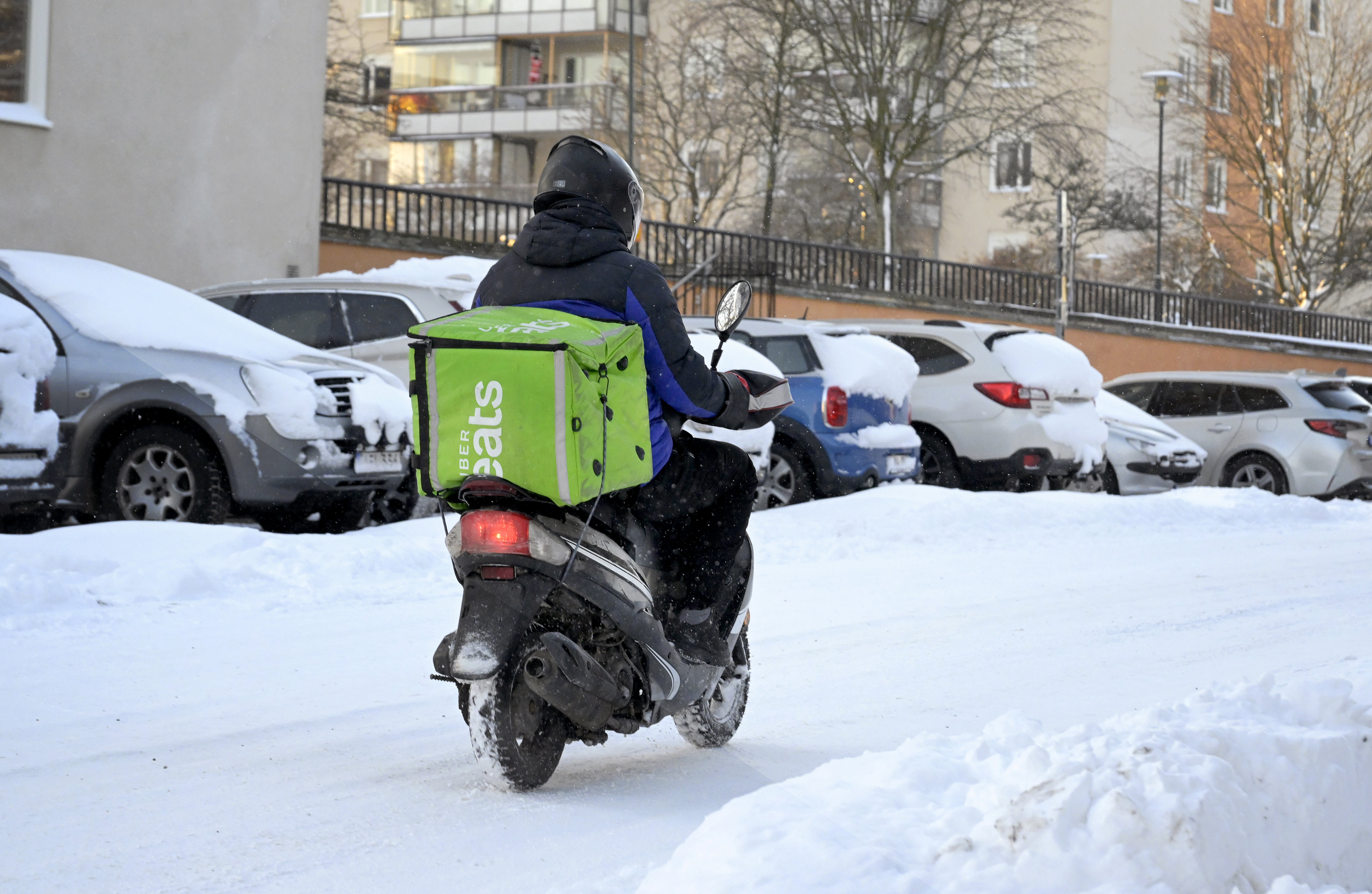 Ett matbud på moped från Uber Eats passerar på en snöfylld gata med parkerade bilar i Solna.