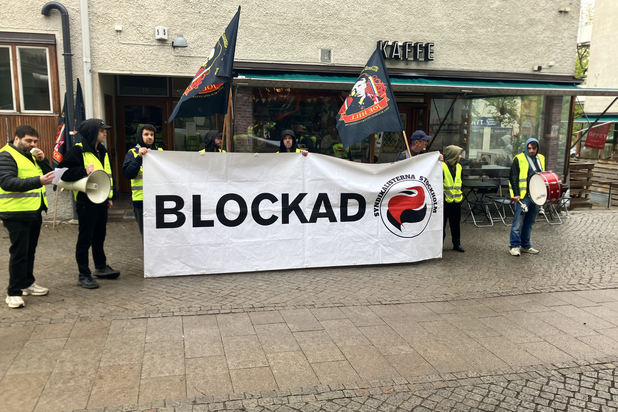 Foto från indrivningsblockad i maj 2026. På bilden står ett 10-tal personer med banderoller och flaggor framför Kaffe Colombia Express Hökarängen 